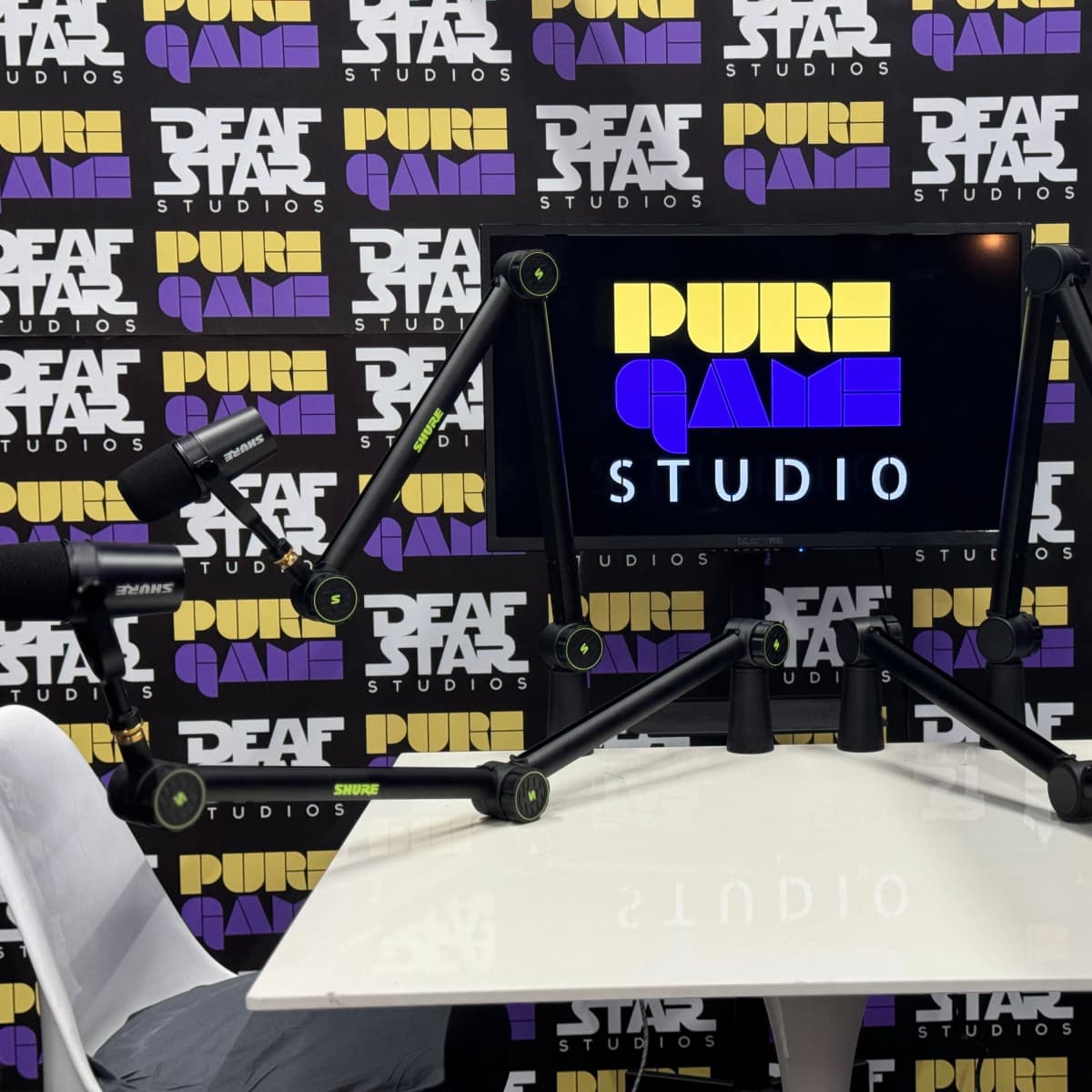 A studio setup with microphones and a monitor displaying "PURE GAME STUDIO" against a patterned backdrop featuring "DEAF STAR STUDIOS."