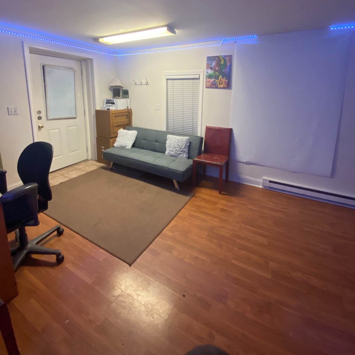 A small room features a gray sofa, a chair, and a light-colored rug, with blue LED lights along the ceiling and a white wall ready for projection.