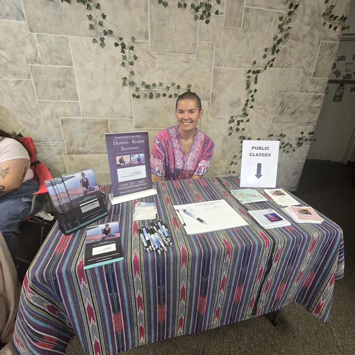 A woman sits at a table covered with a patterned tablecloth, displaying promotional materials for dance classes and a sign indicating public classes.
