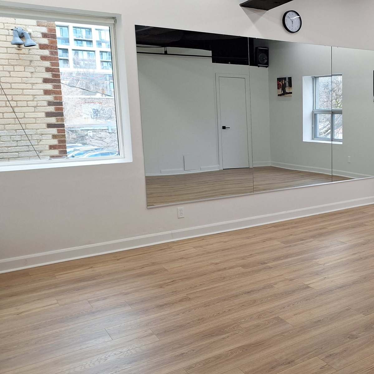 A minimalistic room with wooden flooring, a large mirror on one wall, and two windows providing natural light.