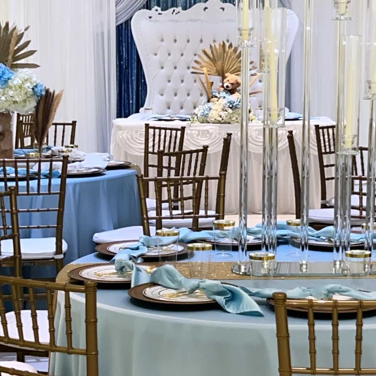 A decorated banquet hall features elegantly set tables with light blue tablecloths, gold chiavari chairs, and a backdrop of a white throne surrounded by floral arrangements and tall candle holders.