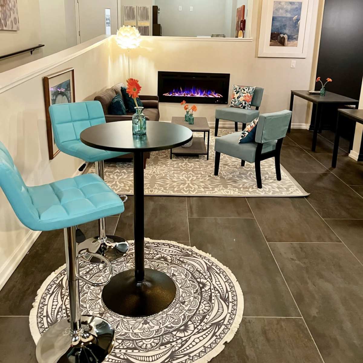 A modern lounge area with turquoise bar stools, a small round table, a fireplace, and decorative artwork on the walls.