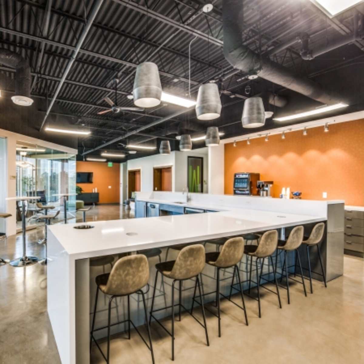 A modern office interior featuring a white countertop bar with seating, glass-paneled meeting rooms, and an orange accent wall.