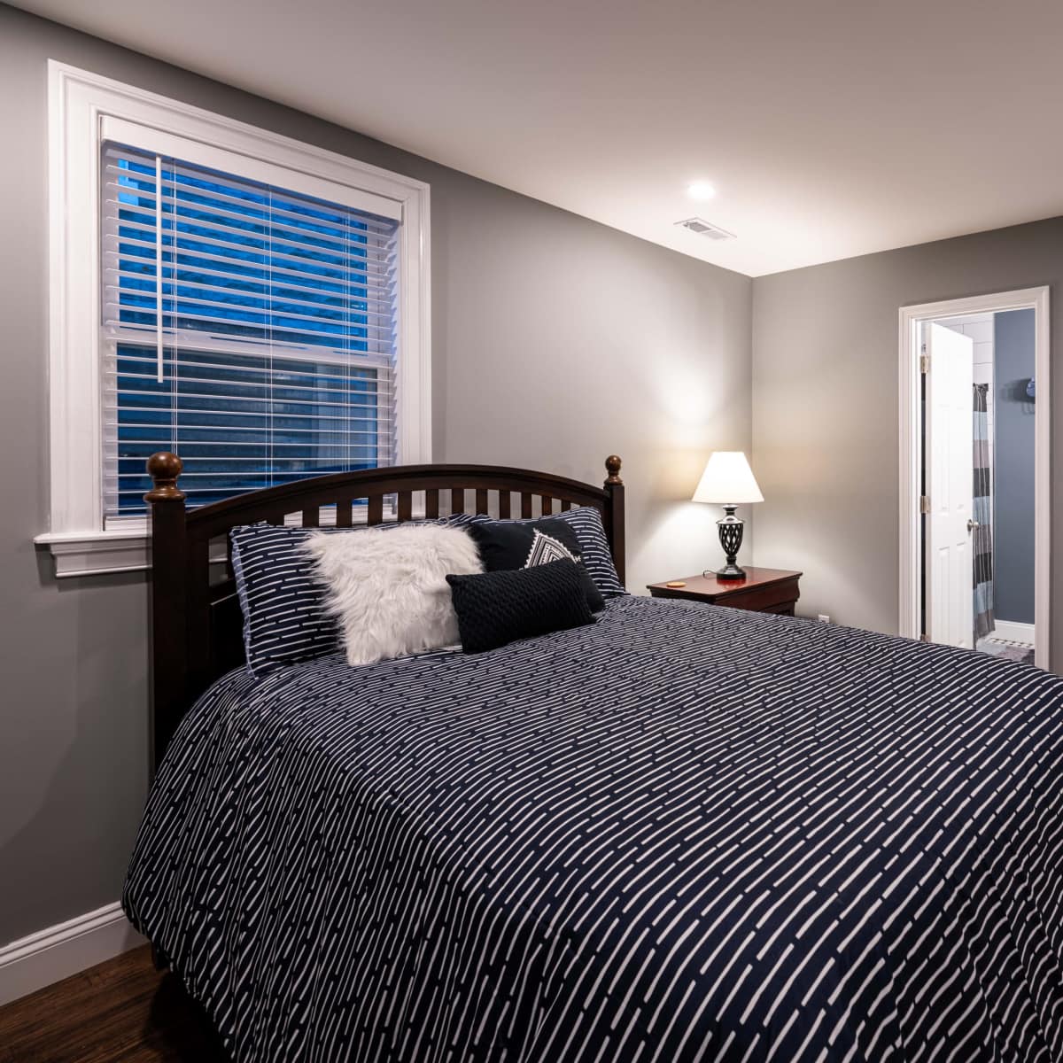 A bedroom with a dark blue patterned bedspread, a wooden bed frame, a nightstand with a lamp, and a door leading to a bathroom.