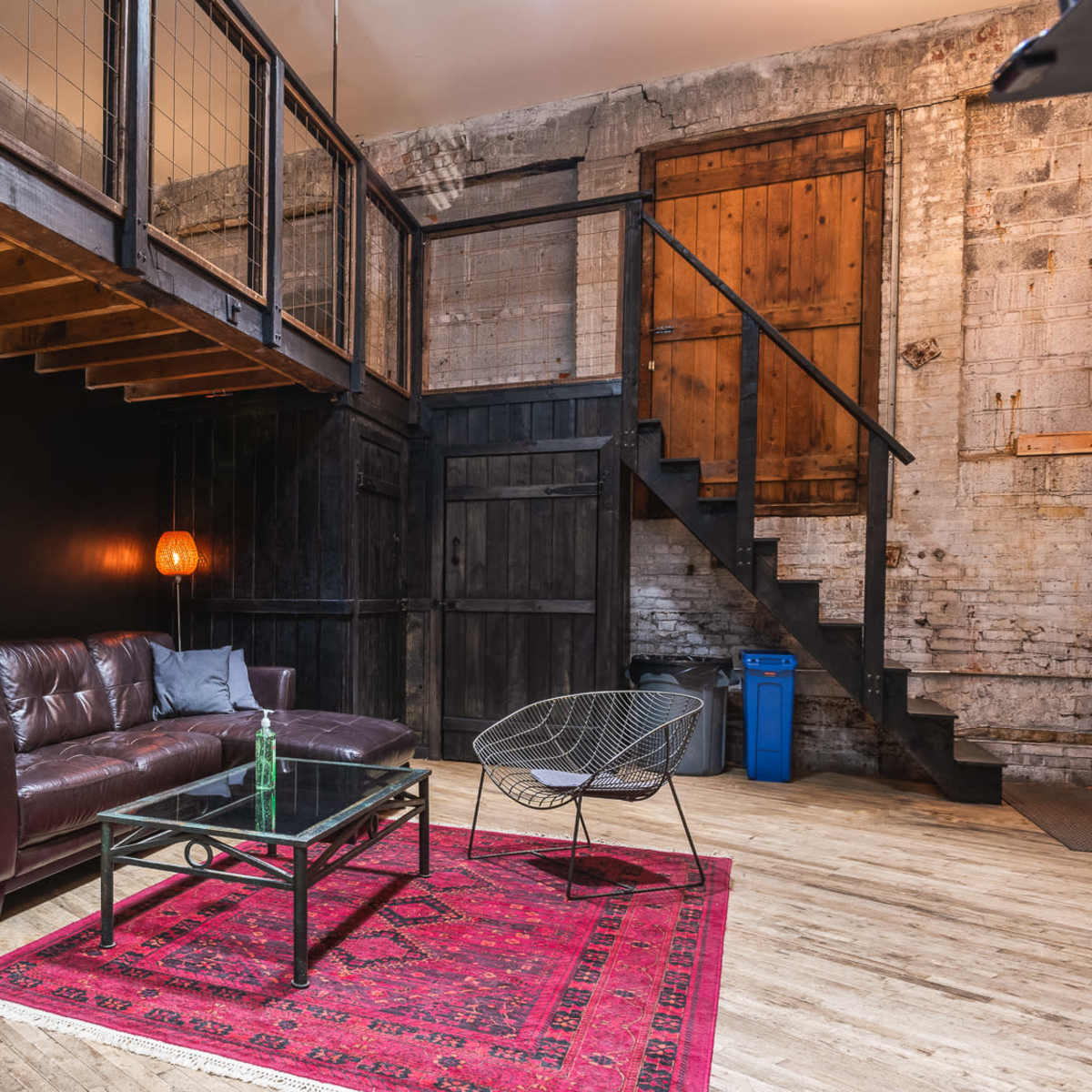 A rustic living space with a dark wooden loft, a leather sofa, a glass coffee table on a patterned rug, and a staircase leading to an upper level.
