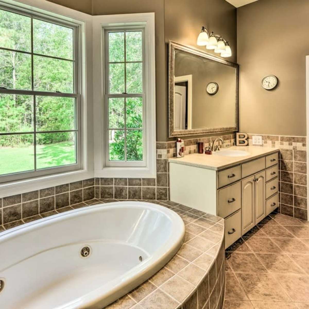 A bathroom featuring a large oval bathtub, a dual sink vanity, large windows overlooking greenery, and tiled walls.