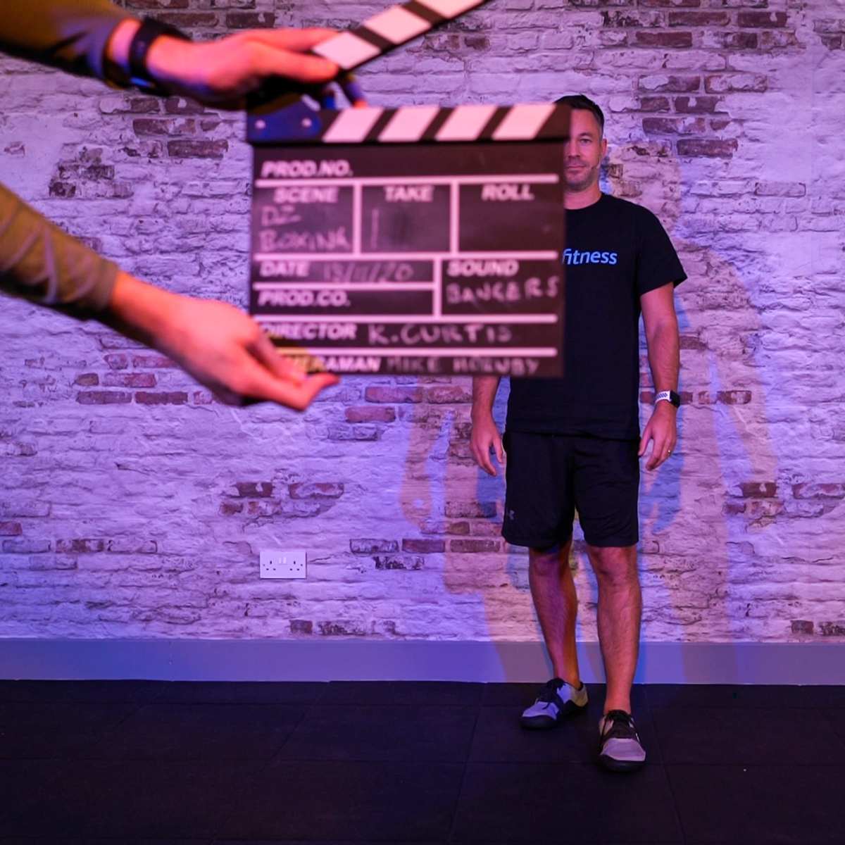 A person stands against a brick wall while another person holds a movie clapperboard in front of them.