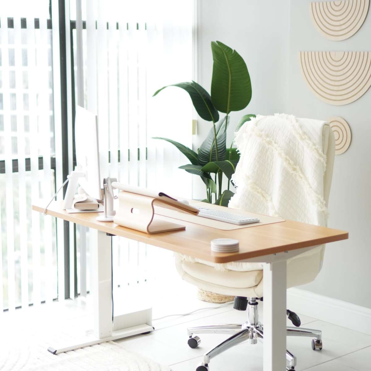 A modern home office features a wooden desk with a computer, a white chair, and a potted plant beside a window with vertical blinds.