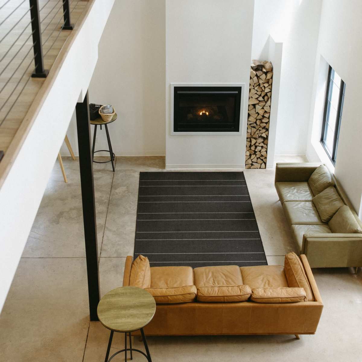 A modern living room with a brown leather sofa, a green couch, a fireplace, and a wooden accent wall, all under a high ceiling with large windows.