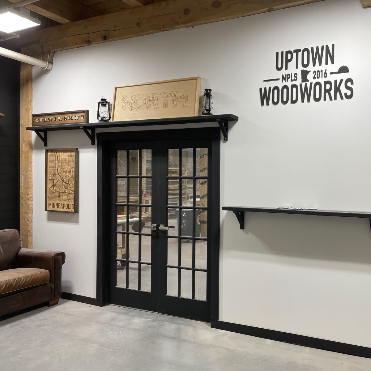 A wall featuring a sign that reads "UPTOWN WOODWORKS" with a framed map and a shelf underneath, alongside double glass doors in a wooden frame.
