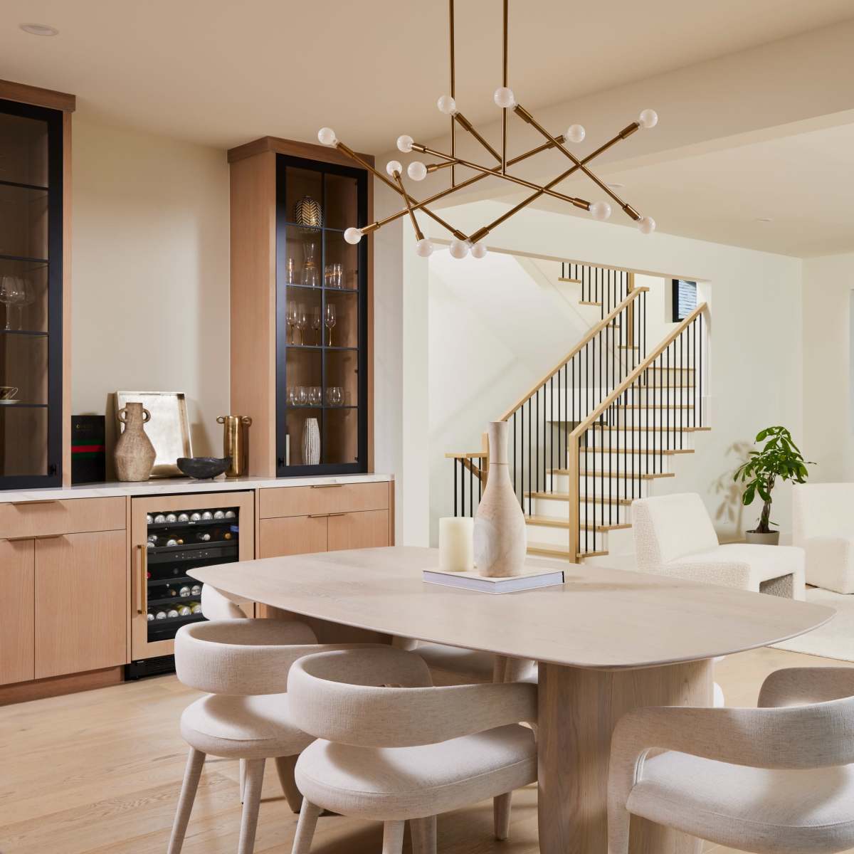 A modern dining area features a wooden table surrounded by curved chairs, with a staircase, cozy seating area, and a fireplace in the background.