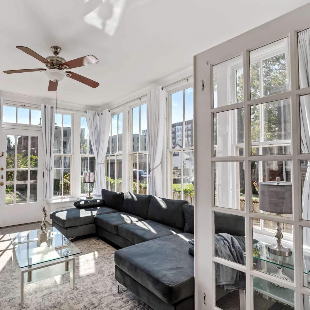 A sunlit sunroom features large windows, a sectional sofa, a glass coffee table, and a ceiling fan.