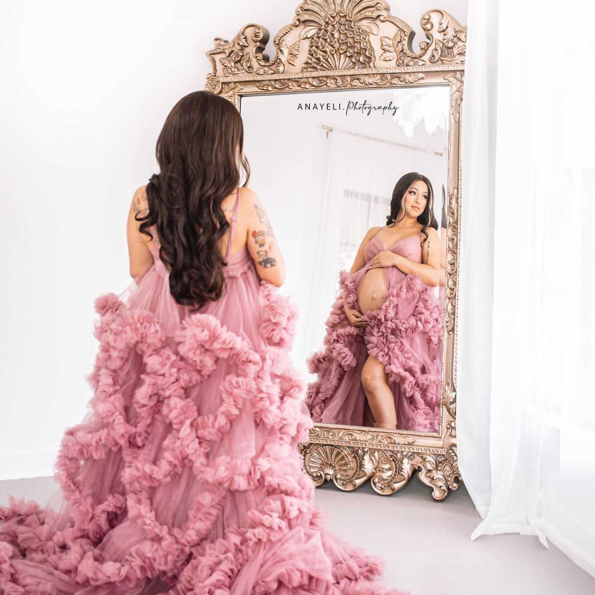 A pregnant woman in a flowing, ruffled pink dress stands in front of an ornate mirror, admiring her reflection.