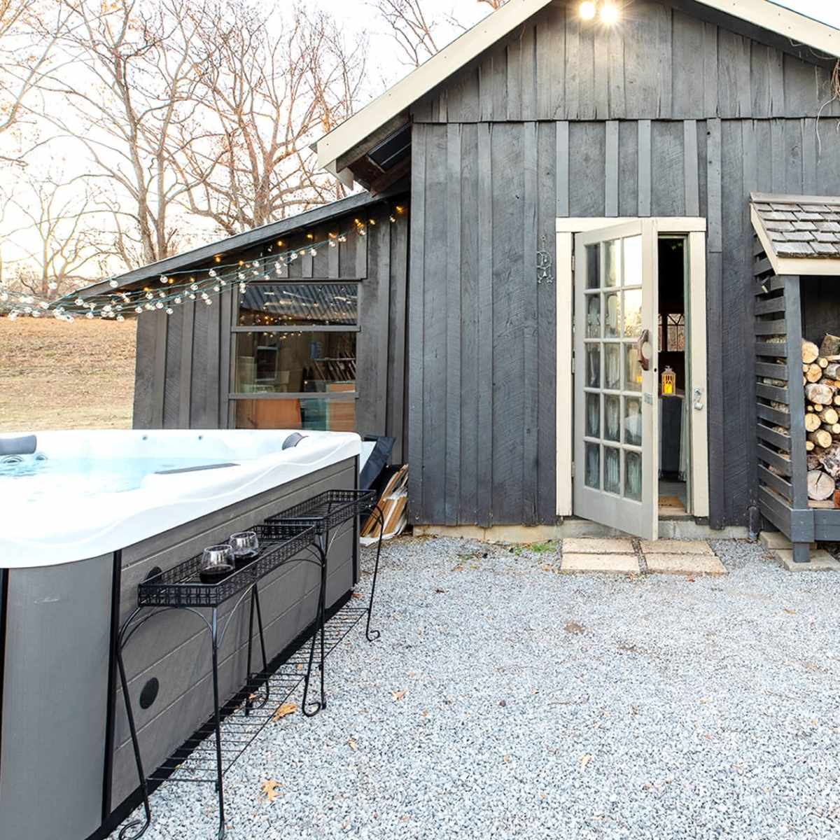 A gray wooden cabin with a hot tub on the left and a stack of firewood against the right side.