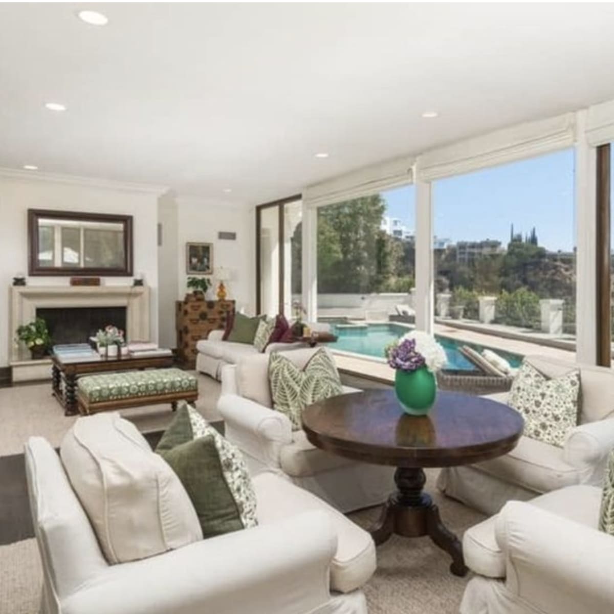 A spacious living room features white sofas arranged around a circular wooden table, with large windows displaying a hillside view.