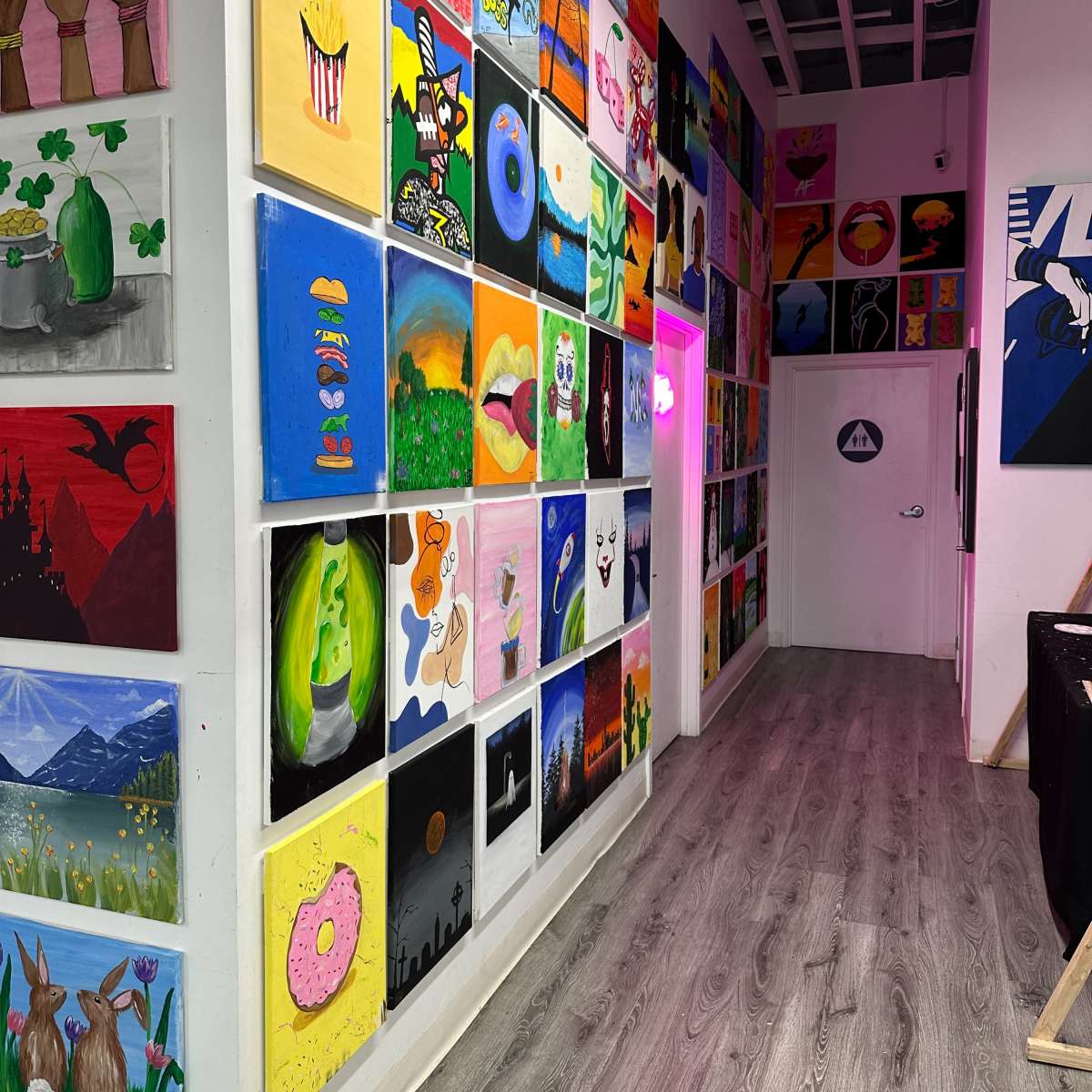 A hallway lined with colorful paintings displayed on white walls, with a door at the end and pink lighting illuminating the space.
