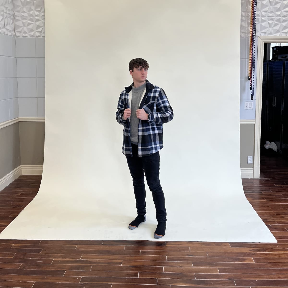 A person in a checkered jacket poses in front of a plain white backdrop in a studio.