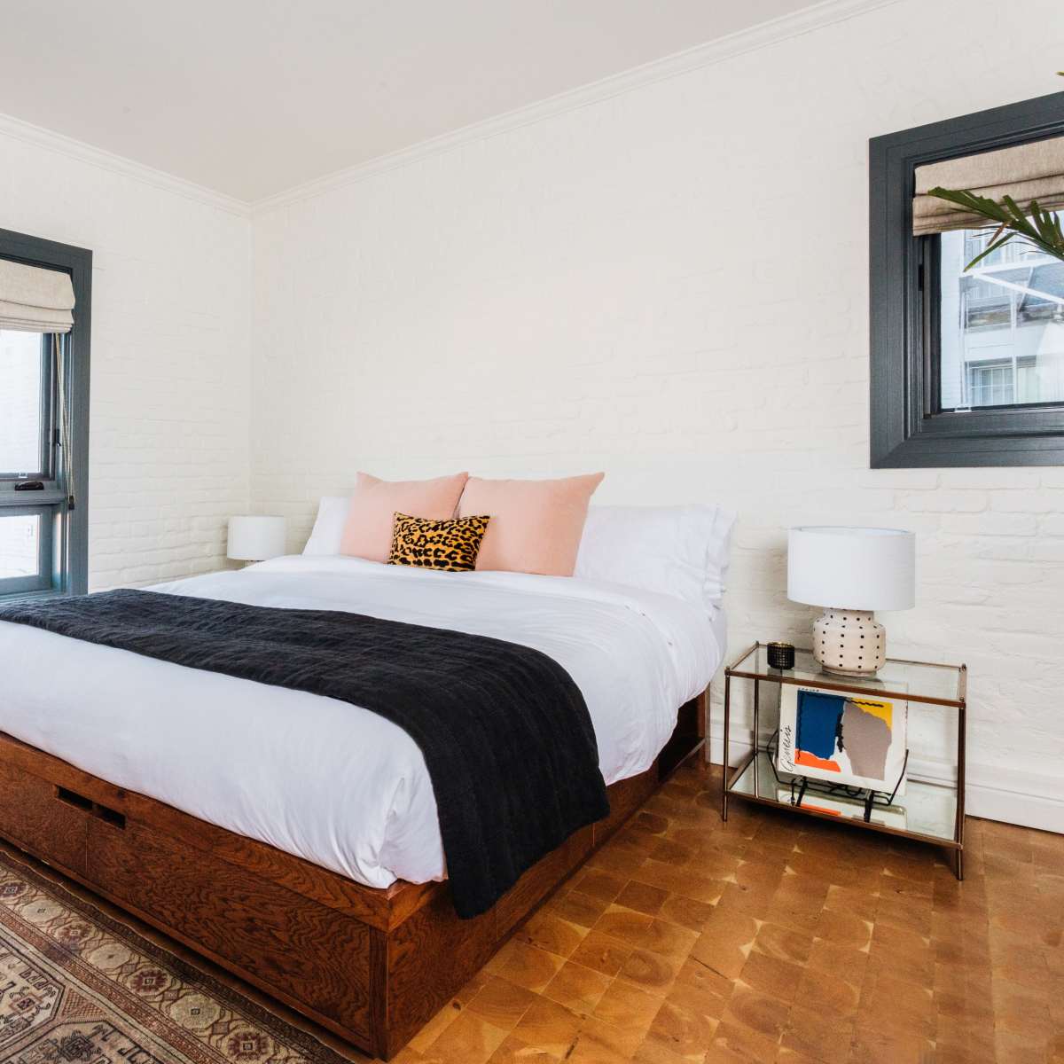 A cozy bedroom featuring a wooden bed with white bedding, a patterned rug, a nightstand with a lamp, and large windows with blinds.