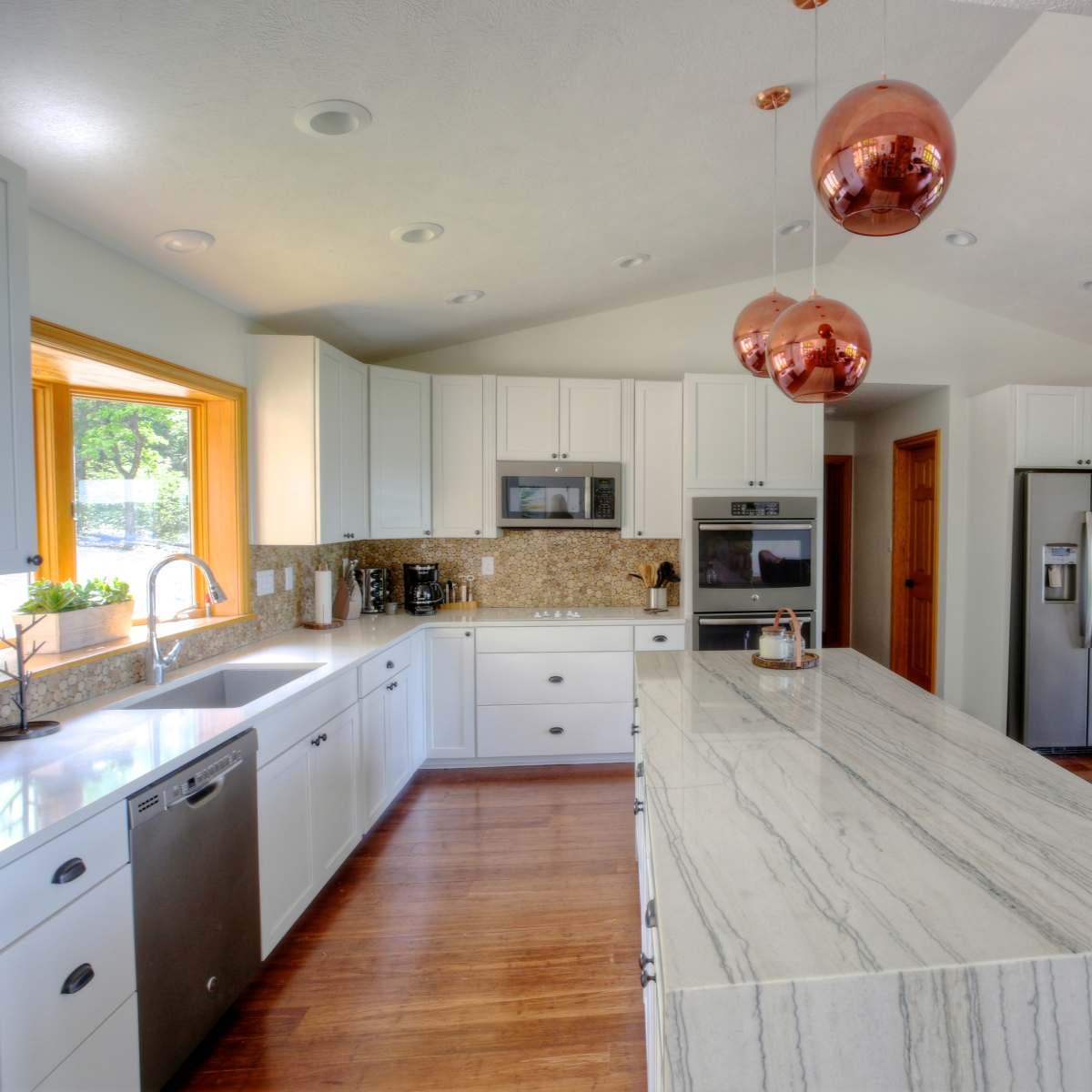 A modern kitchen with white cabinets, a large marble island, and copper pendant lights, leading to a dining area with a round table and chairs.