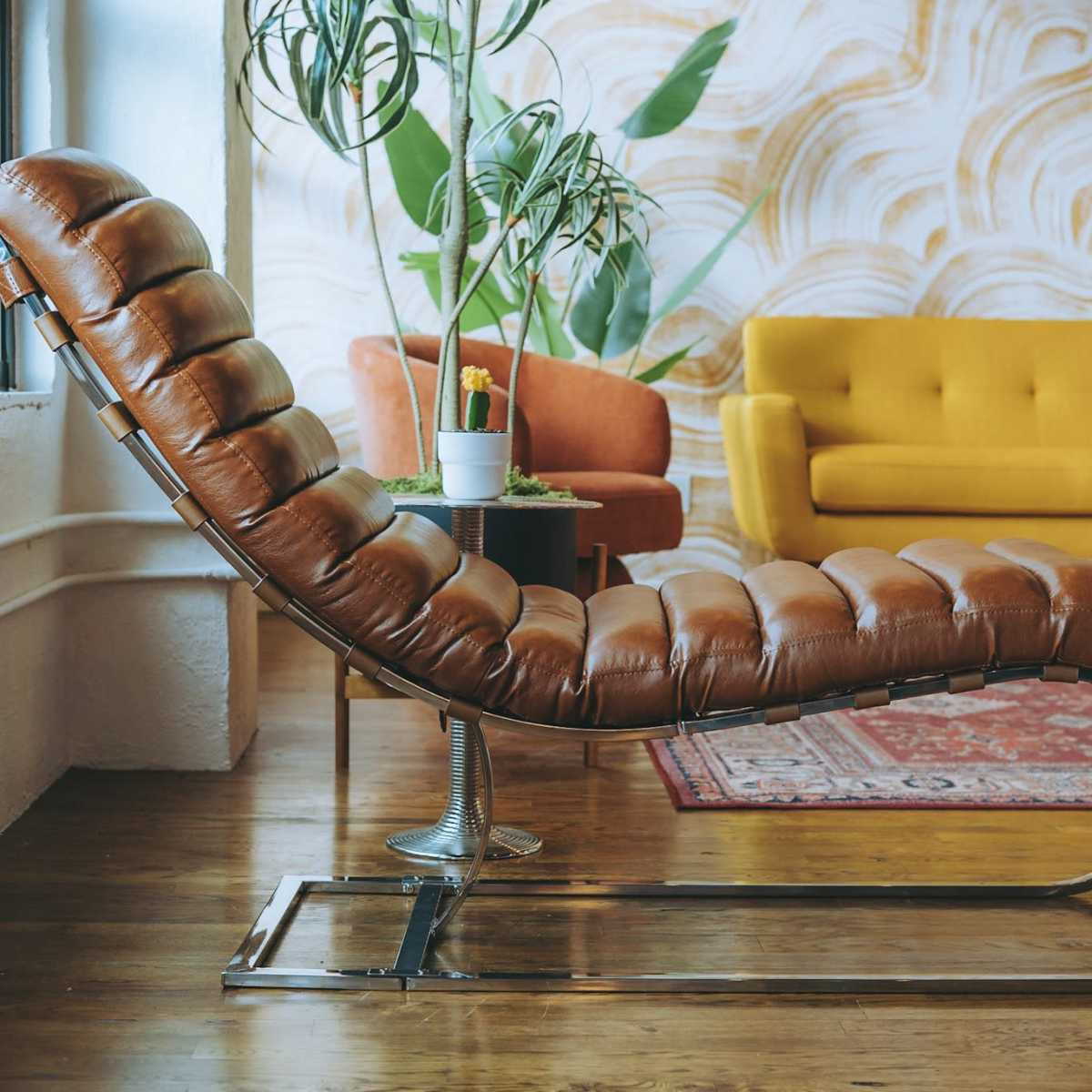 A brown leather chaise lounge is positioned beside a yellow sofa in a room with a patterned wall and wooden flooring.