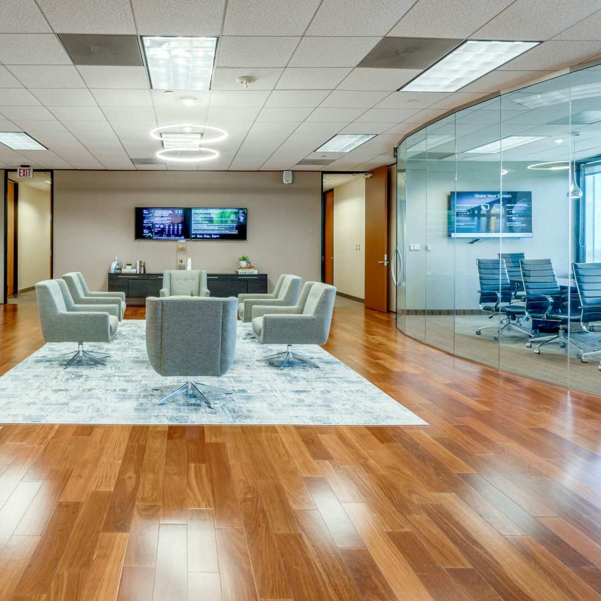 A modern office space with gray armchairs arranged around a circular rug, flanked by tall potted plants and a glass-walled meeting room in the background.