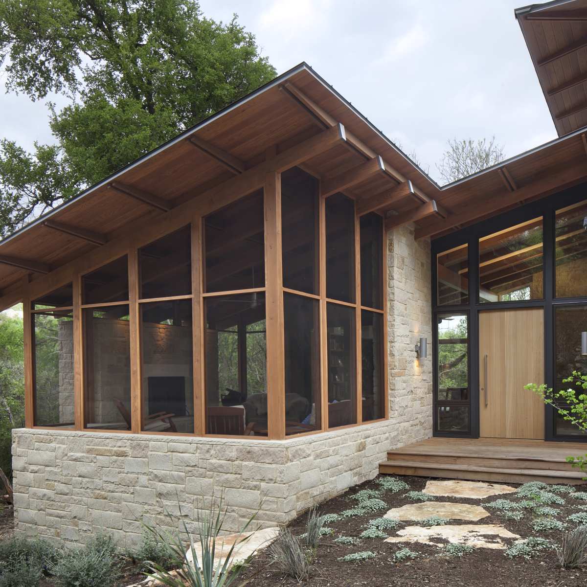 A modern house with a stone exterior and large glass windows, featuring a sloped wooden roof and a landscaped entrance.