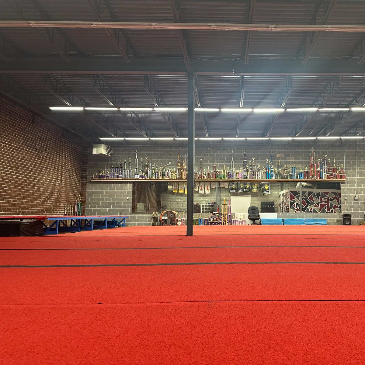 A gymnasium with a red floor, lined with trophies on shelves against a brick wall, and blue mats in the center.