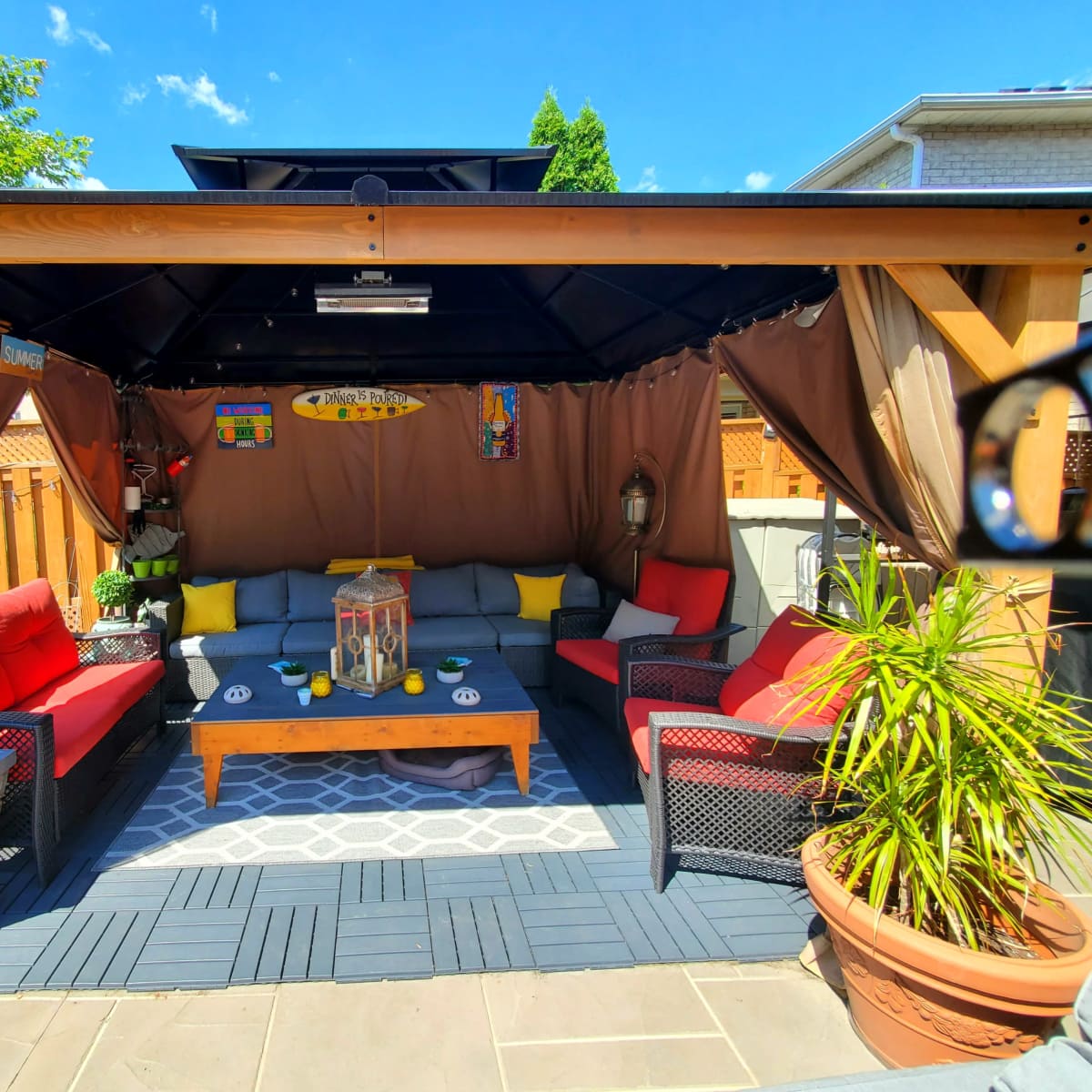 A covered outdoor seating area features red and yellow cushions on black wicker furniture, a wooden coffee table, and potted plants under a tent-like structure.