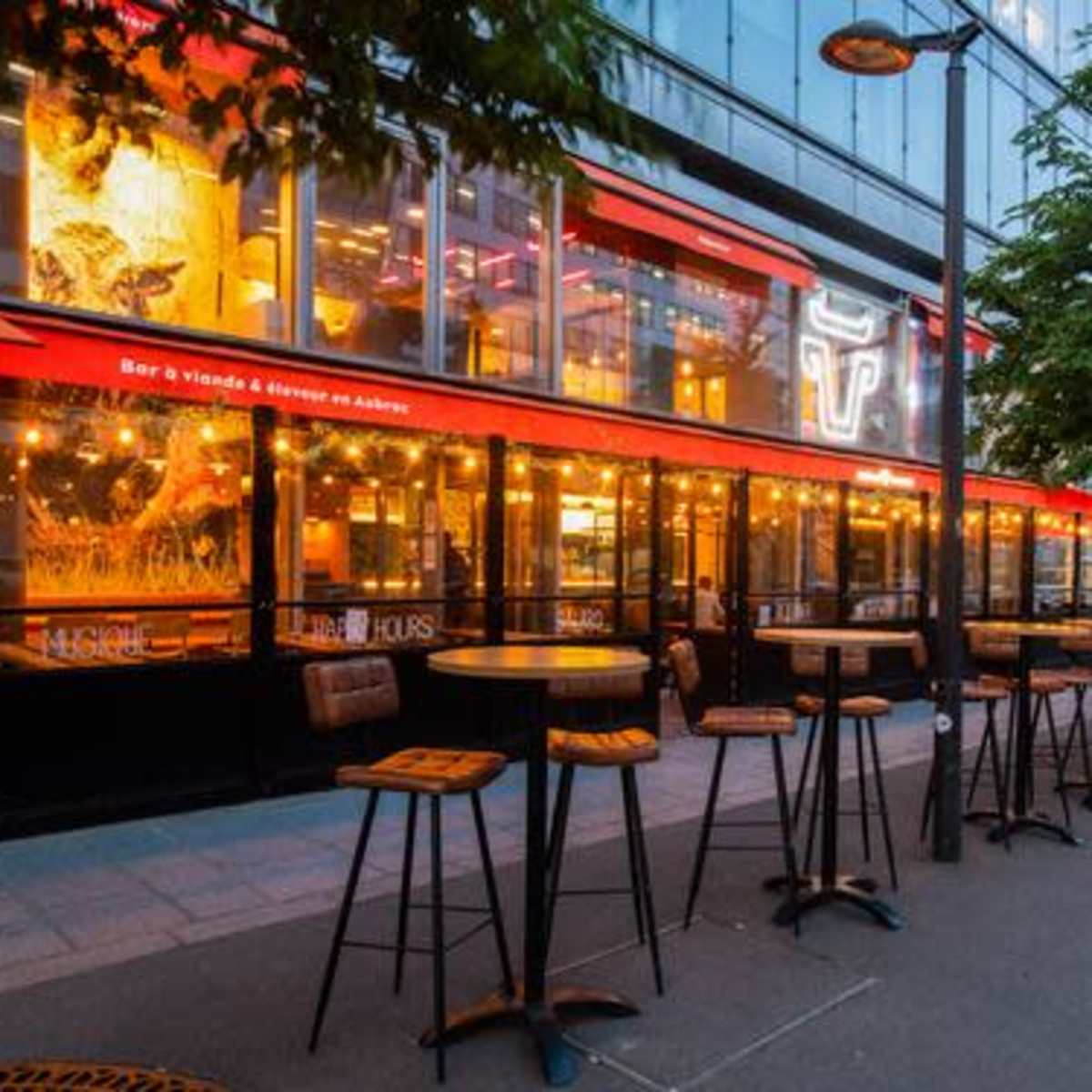 An outdoor seating area of a restaurant with high tables and chairs along a walkway, featuring large windows and a red awning.