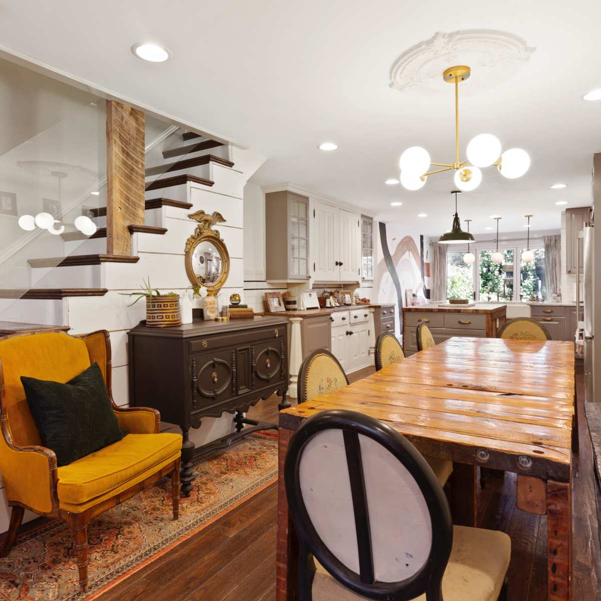 A modern kitchen and dining area featuring a wooden dining table, an ornate light fixture, and a staircase with a glass railing.