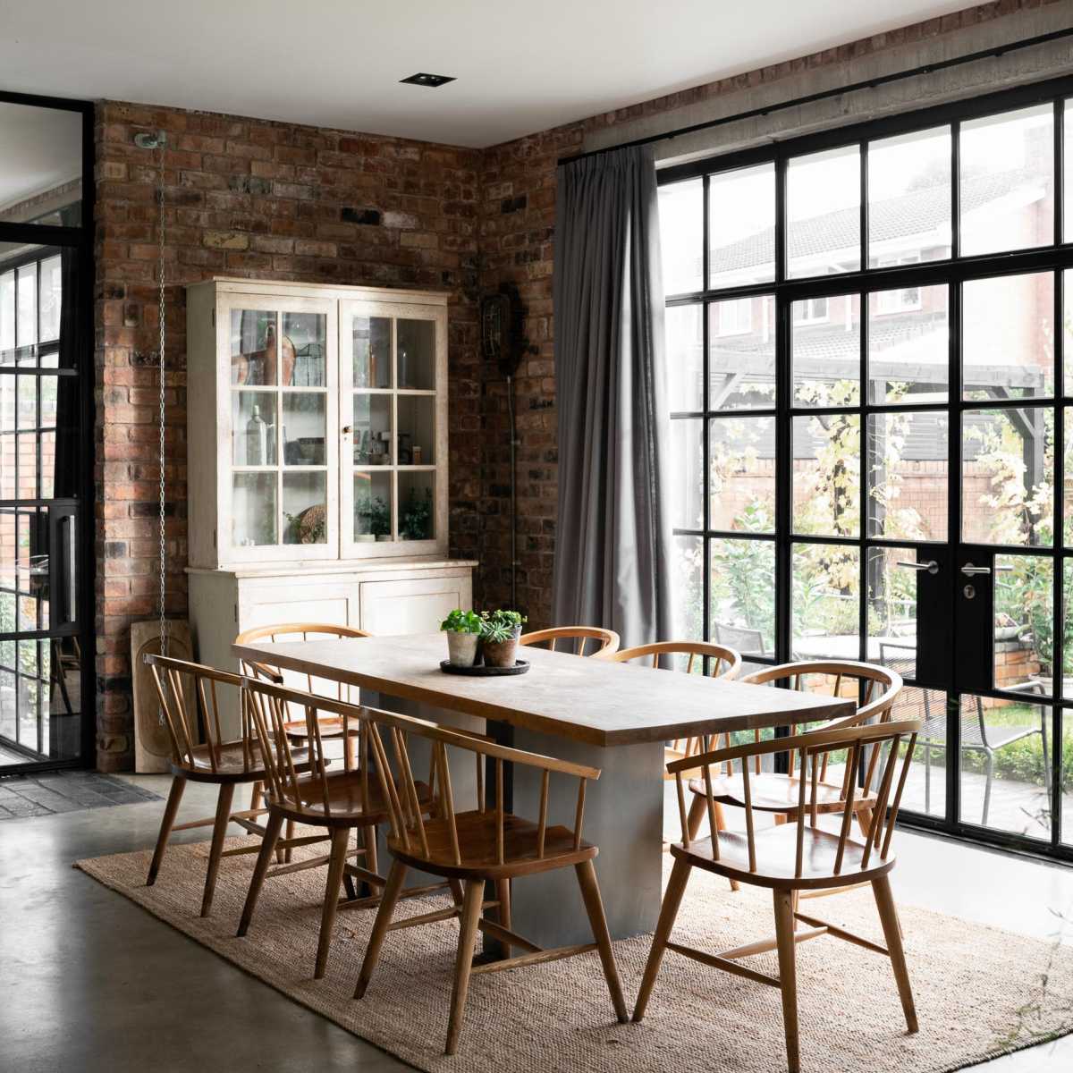 A modern dining area features a long table surrounded by wooden chairs, with large windows that overlook a garden.
