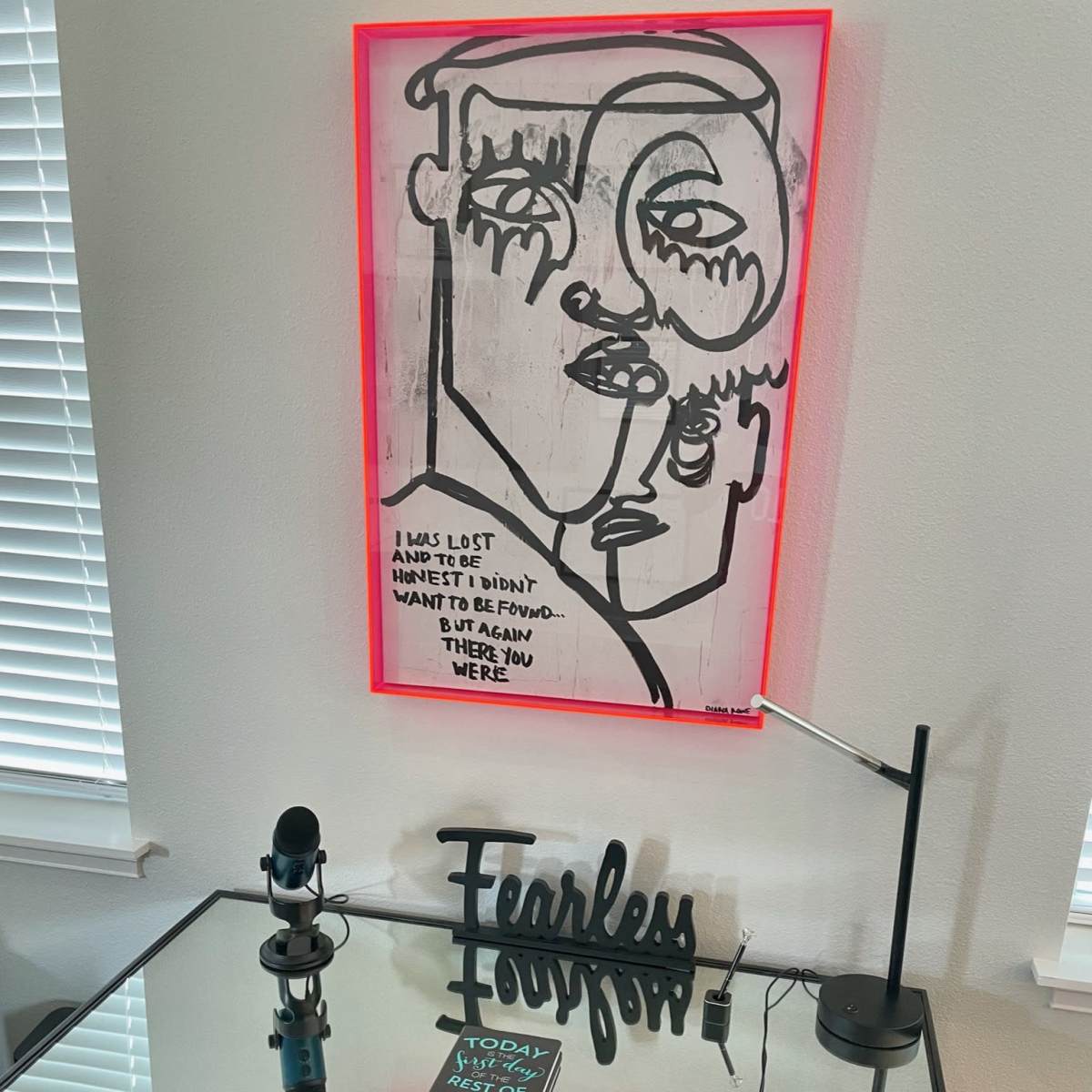 A modern desk with a glass top, featuring a decorative sign that reads "Fearless Forever," a book titled "Today Is Your Day," a microphone, and a neon-lit artwork on the wall depicting abstract faces.