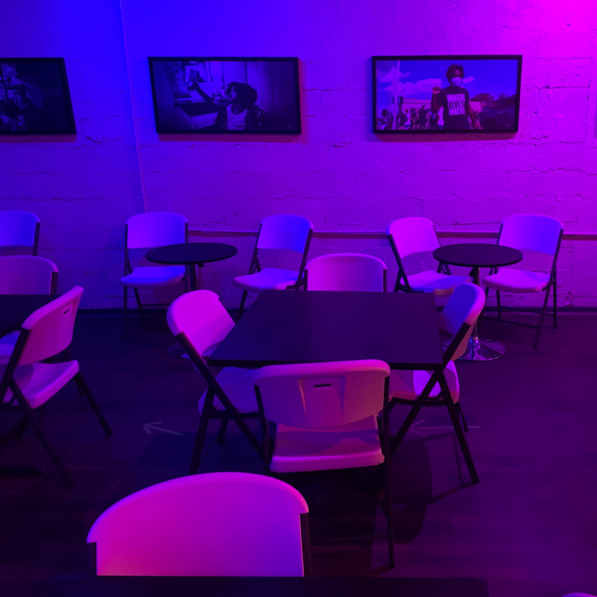 A dimly lit café features several black tables and white folding chairs against a wall adorned with framed photographs.