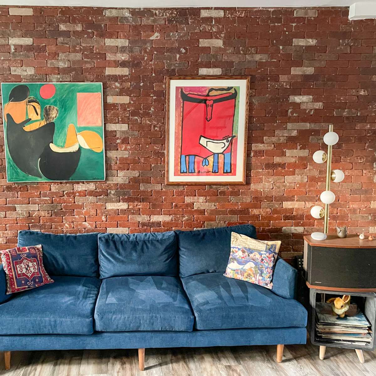 A blue couch in front of a brick wall, adorned with two colorful paintings and flanked by stylish side tables with decorative items.