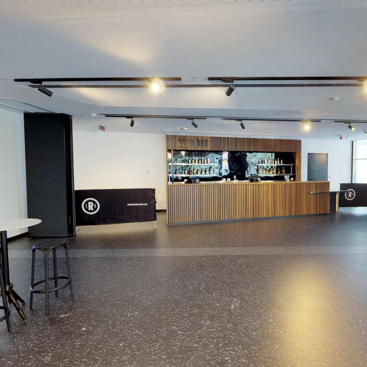 A modern bar area with a wooden counter, shelves behind it filled with bottles, and a seating area featuring a black couch and high chairs.