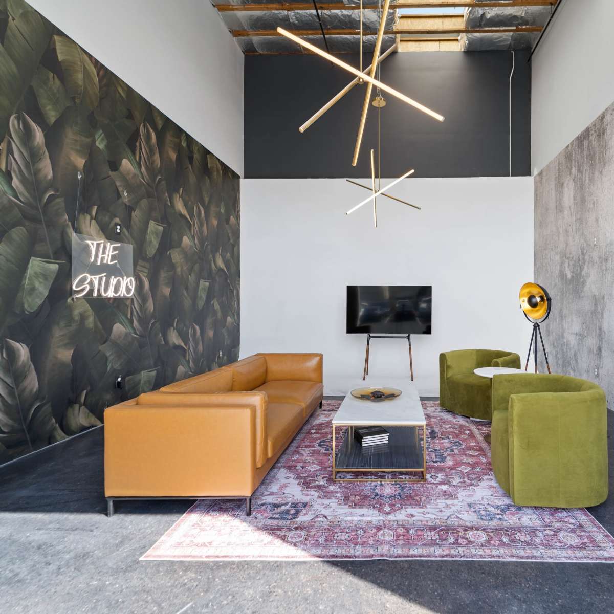 A modern studio space with a brown leather sofa, green accent chairs, a patterned rug, and a television mounted on the wall, set against a backdrop of botanical and concrete textures.