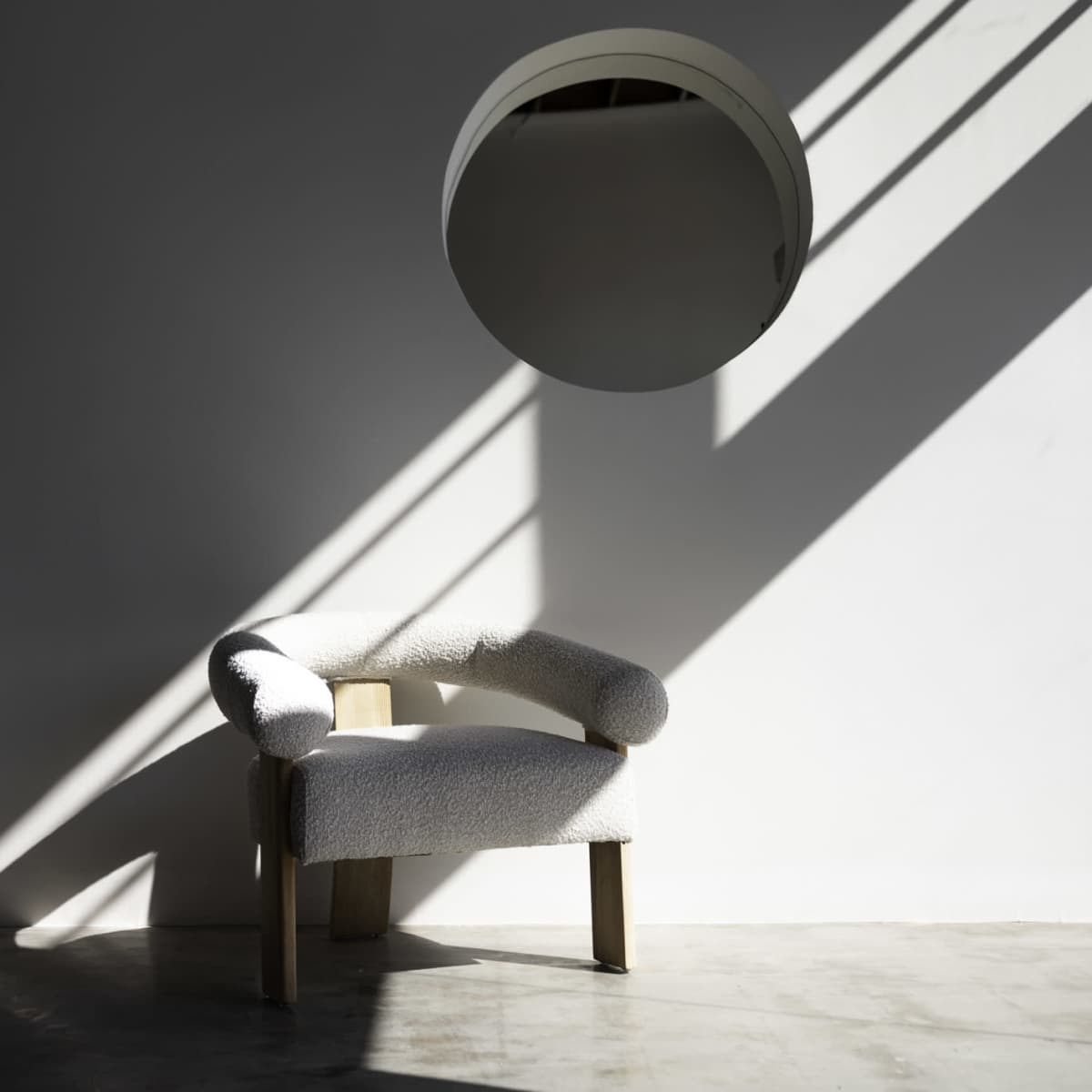 A textured chair sits against a wall, illuminated by angled shadows and accompanied by a round mirror.