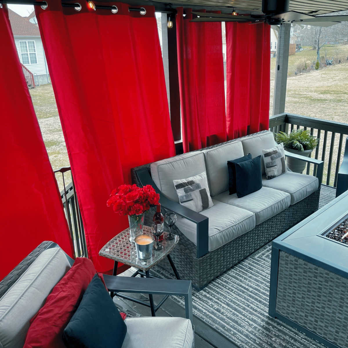 A covered outdoor patio features a seating area with a fire table, surrounded by red curtains and decorated with flowers and cushions.