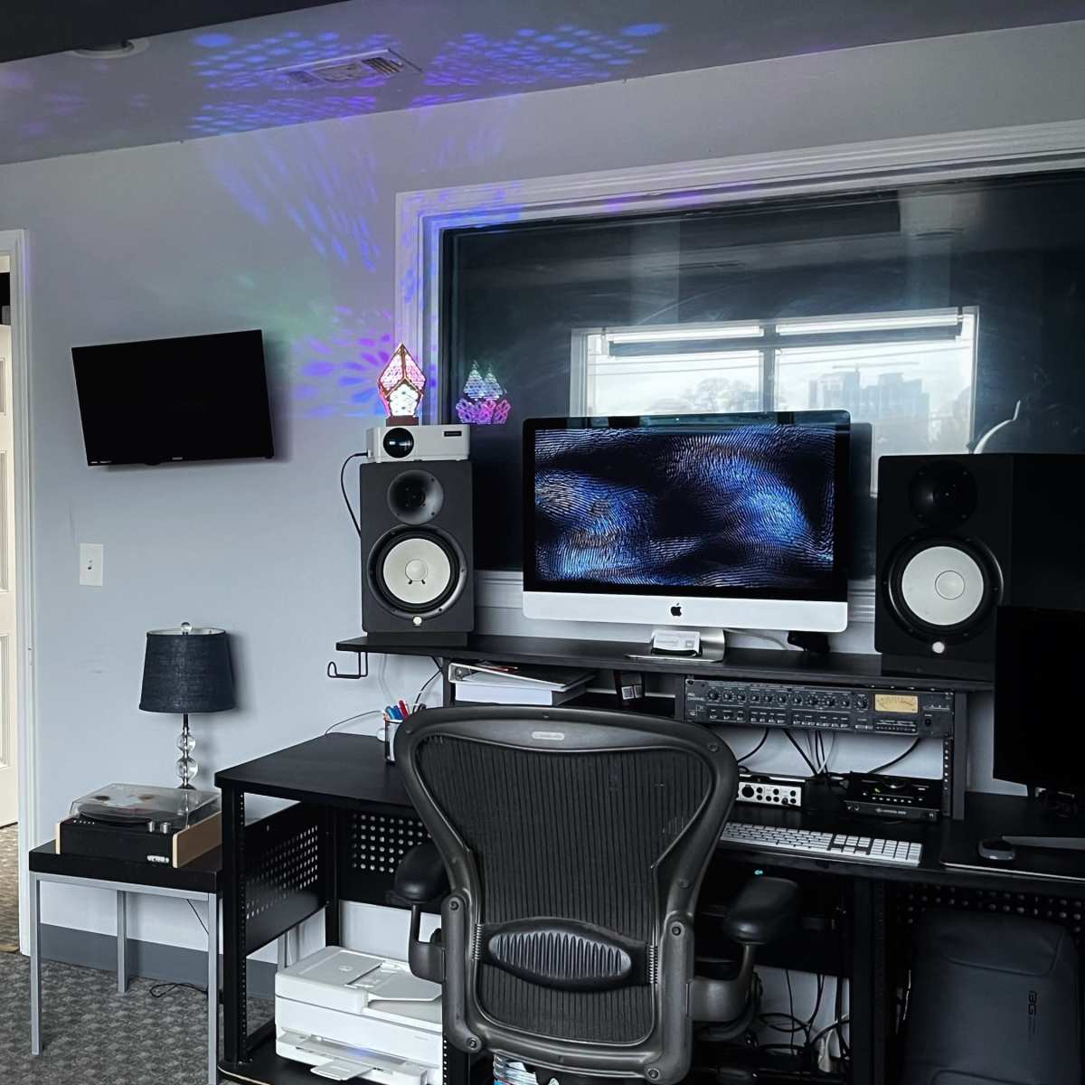 A music studio features a desk with audio equipment, a computer monitor, a chair, and a doorway leading to another room.