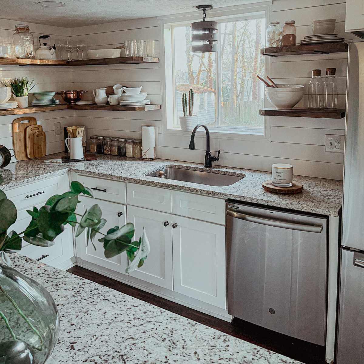 A modern kitchen features white cabinetry, open shelves with various dishware, and a window overlooking greenery.