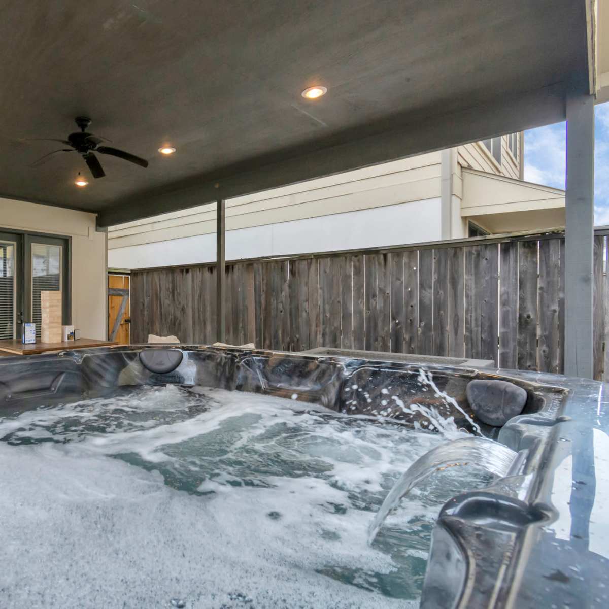 A hot tub with bubbles under a covered patio, adjacent to a wooden fence and a sliding glass door.