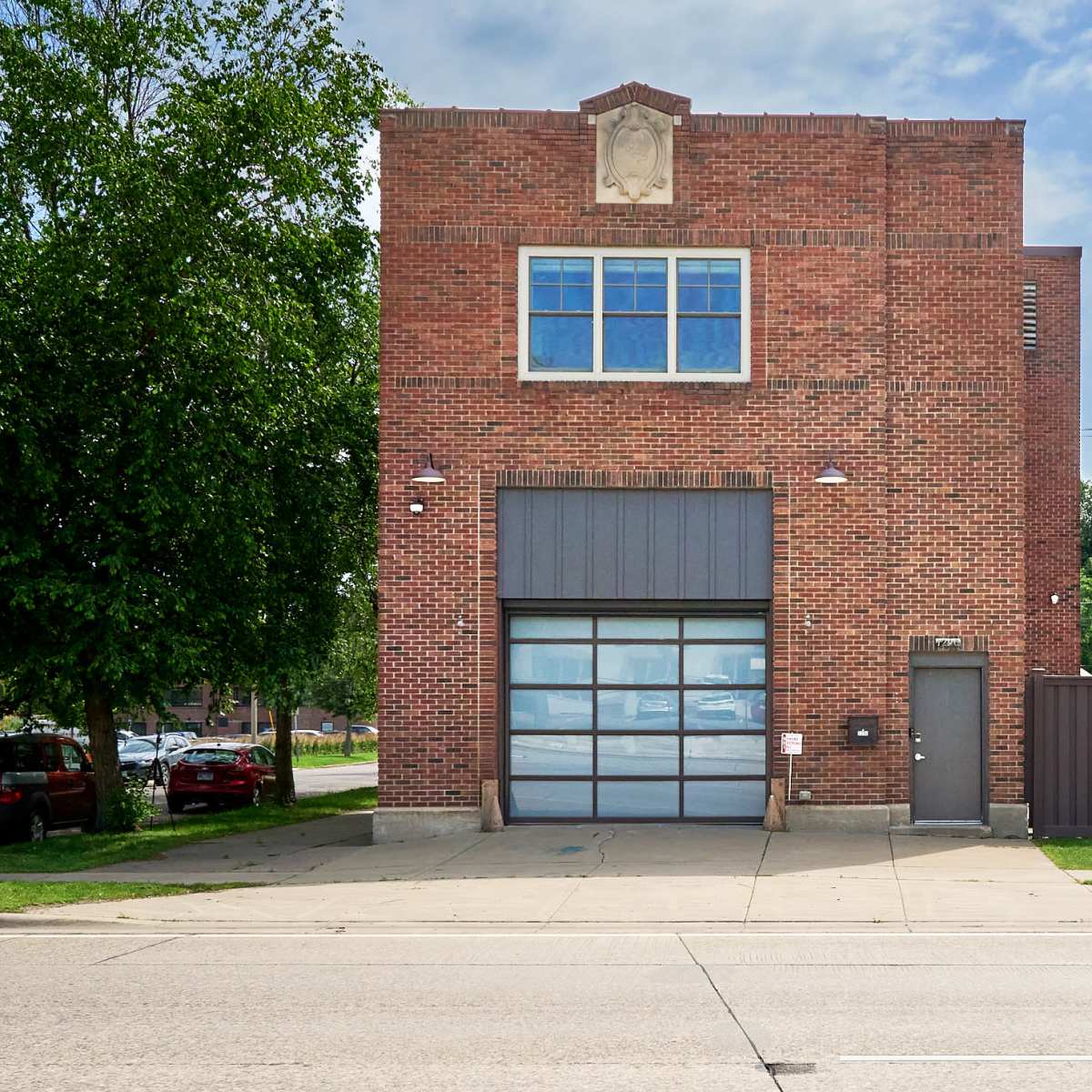 A two-story brick building with large windows and a garage door, situated on a paved street with green grass and a fence alongside it.