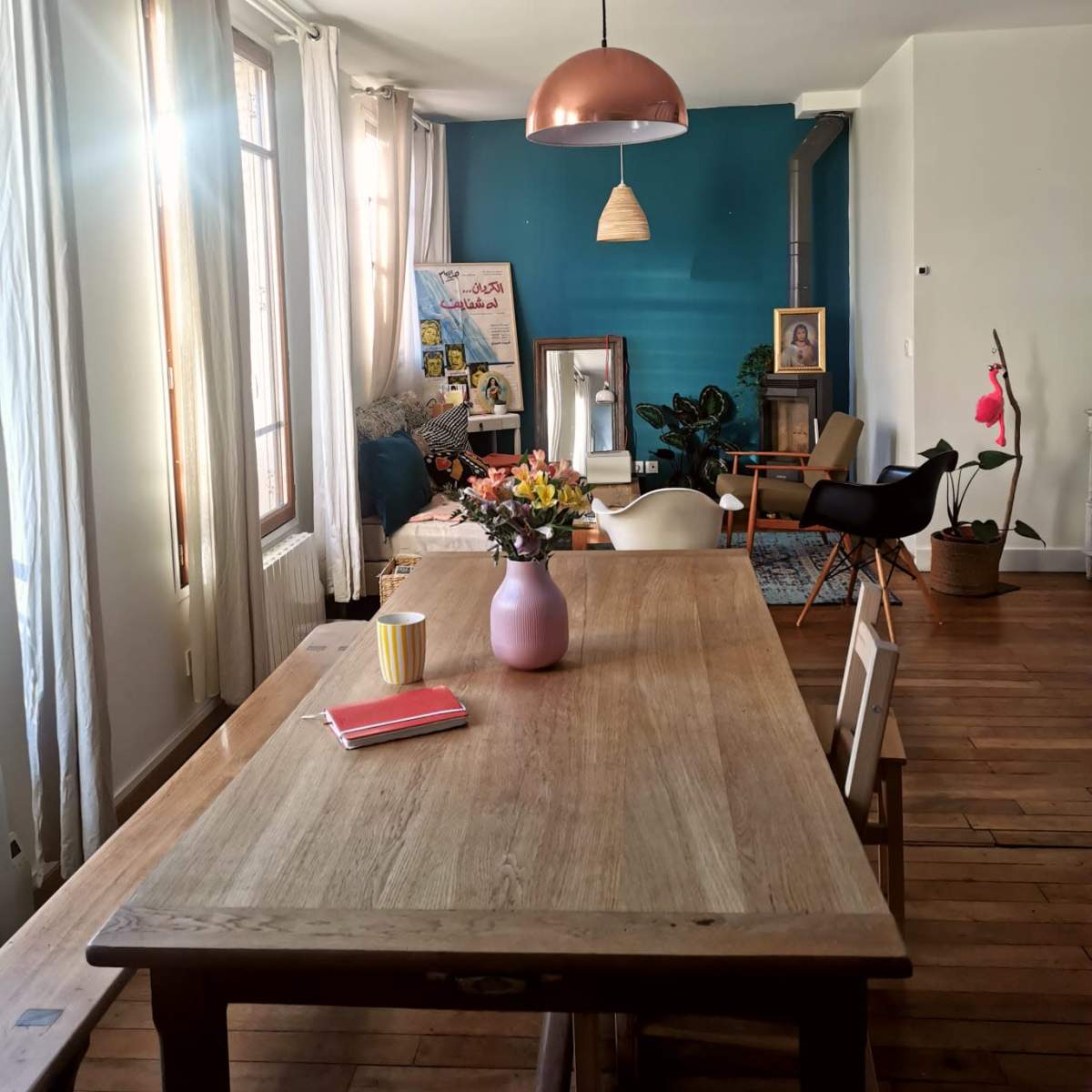 A dining area features a wooden table with a pink vase of flowers, booklets, and a yellow cup under a copper pendant light, with a blue accent wall and chairs in the background.