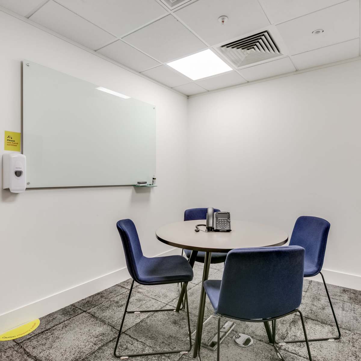 A small meeting room featuring a round table with four blue chairs, a whiteboard, a wall-mounted TV, a phone, and a hand sanitizer dispenser.