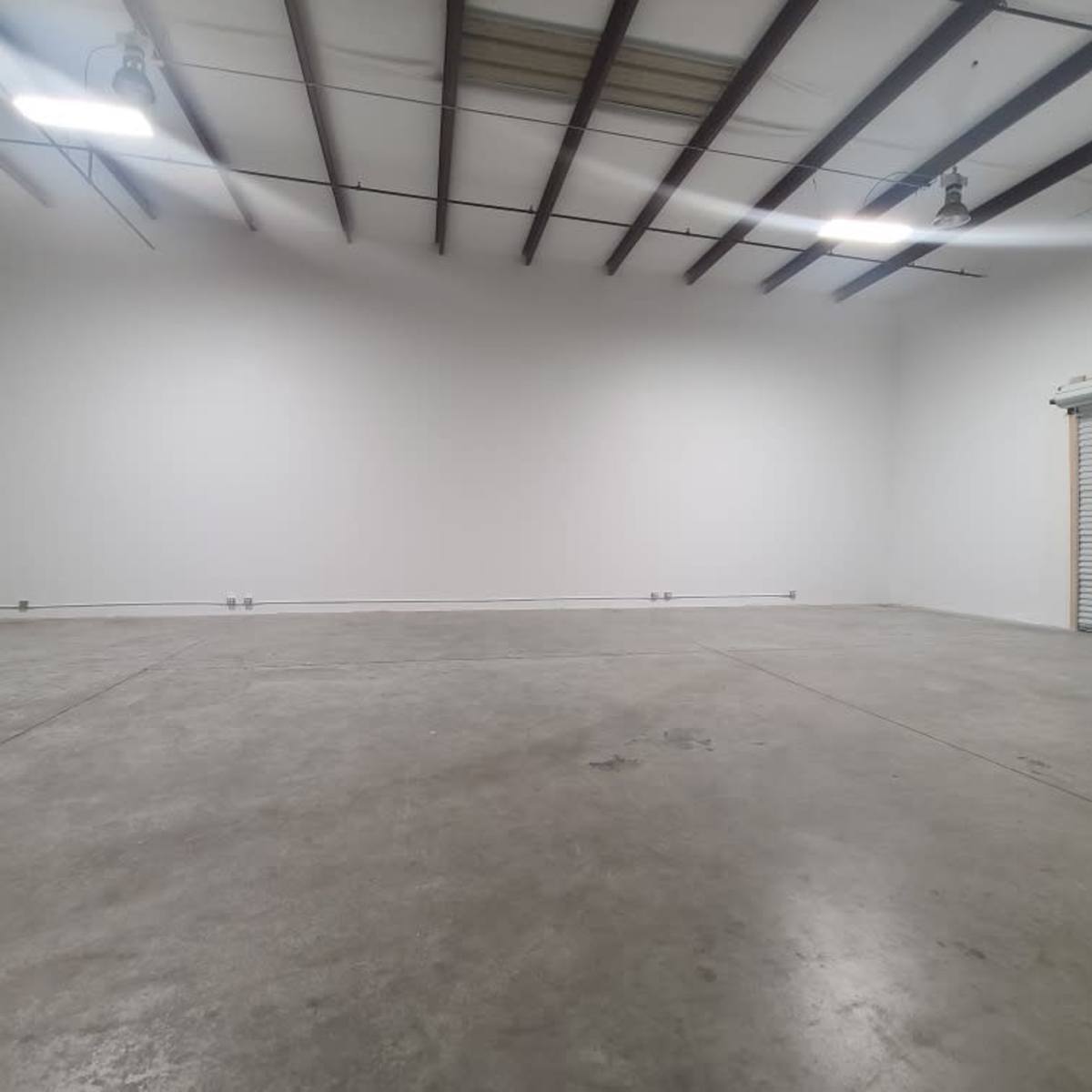 A spacious, empty industrial interior with concrete flooring, high ceilings, and a roll-up garage door.