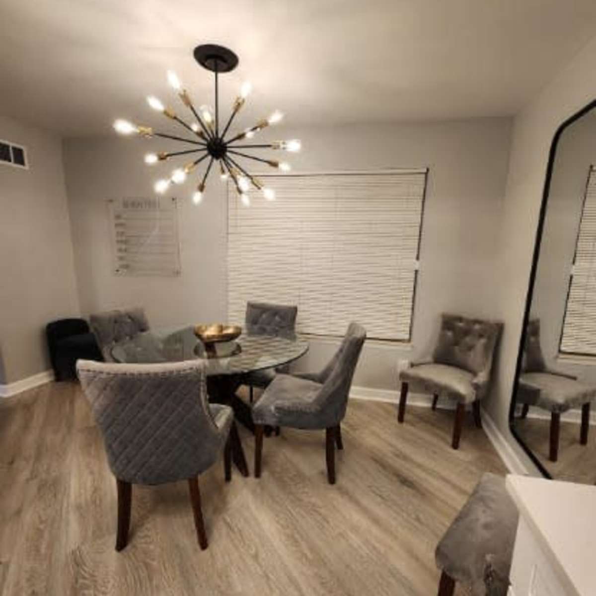 A modern dining area featuring a glass table surrounded by six upholstered chairs, with a large chandelier overhead and a mirrored wall.
