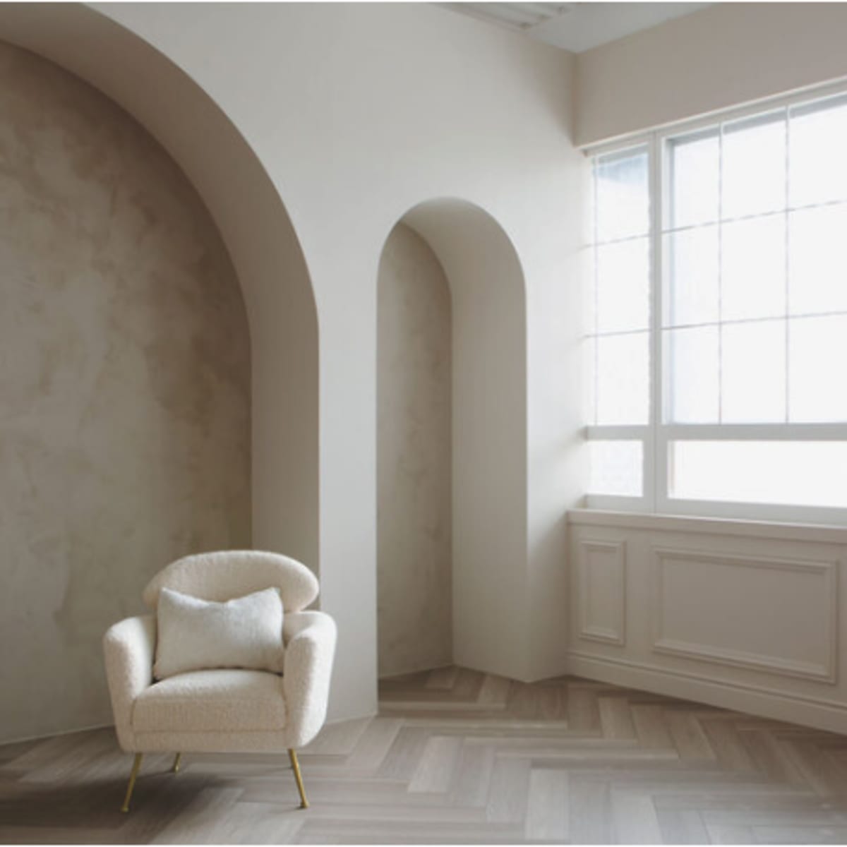 A light-filled room features a minimalist upholstered chair positioned against a textured wall and arched alcove near a large window.