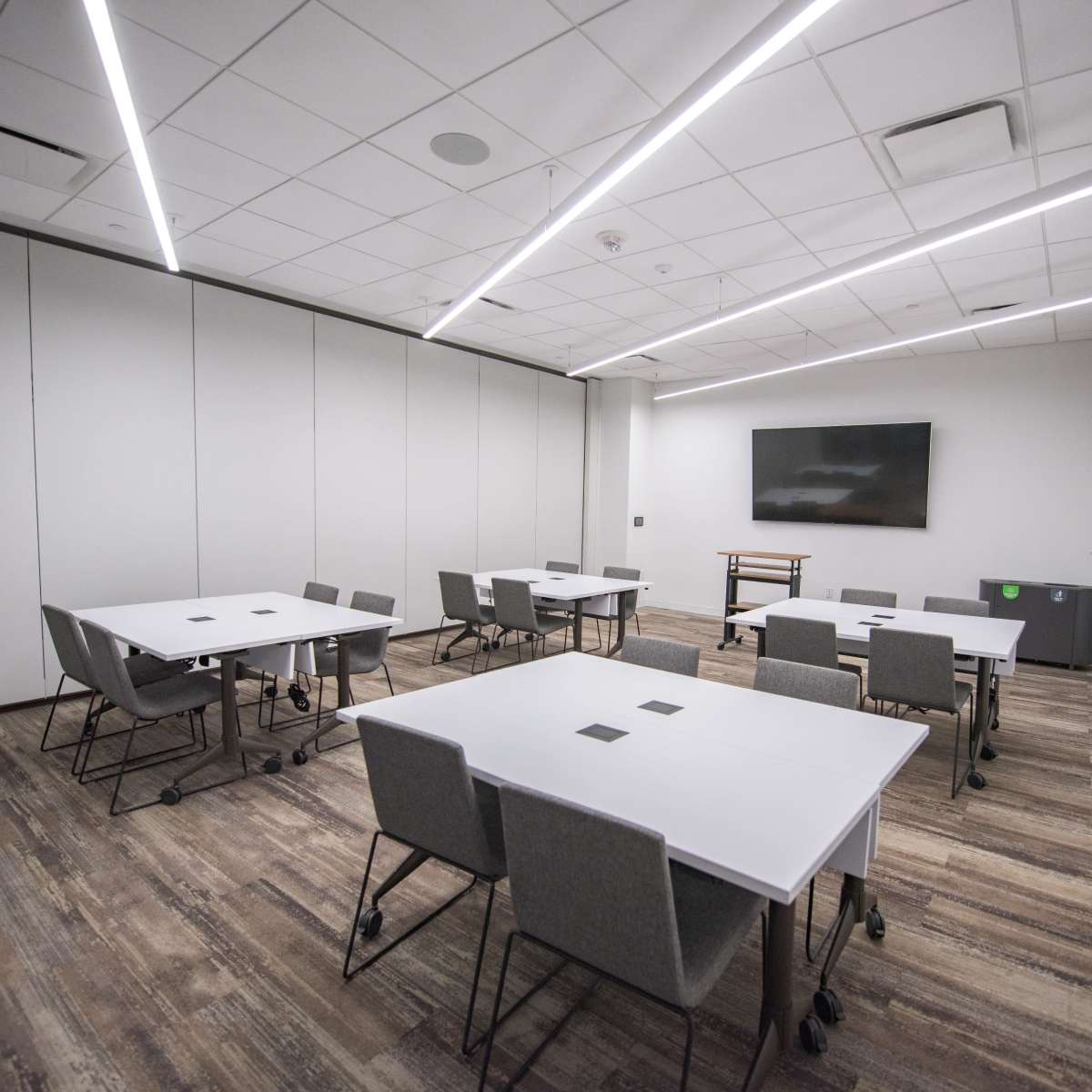 A modern conference room features multiple white tables and gray chairs arranged around the space, with a wall-mounted screen at one end.