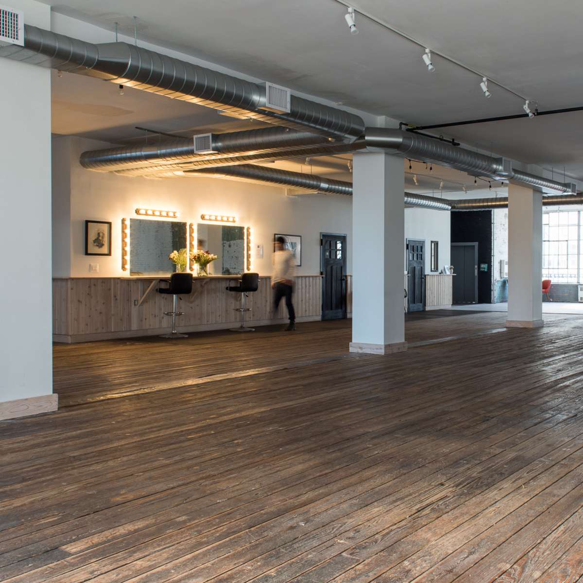 A spacious interior with wooden floors, large windows, and minimalist decor featuring a few furnishings and fixtures along the walls.