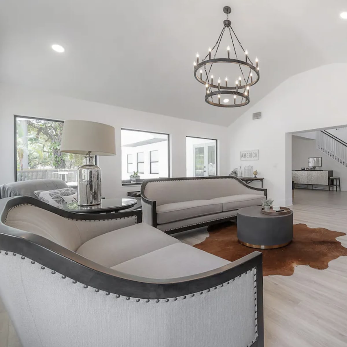 A spacious living room with two upholstered sofas, a round coffee table on a cowhide rug, and large windows allowing natural light to enter.